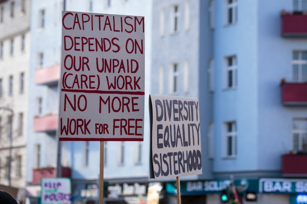 Image of protestors with sign that reads Capitalism depends on unpaid care work. No more Work for Free.