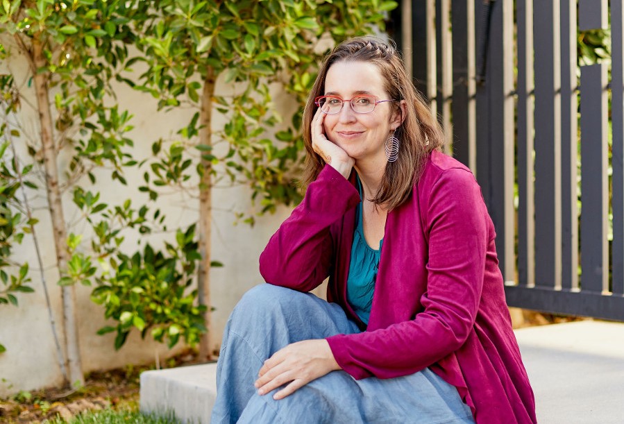 A mature white woman with brown hair and glasses sits in a backyard, smiling and cupping her head in her hand.