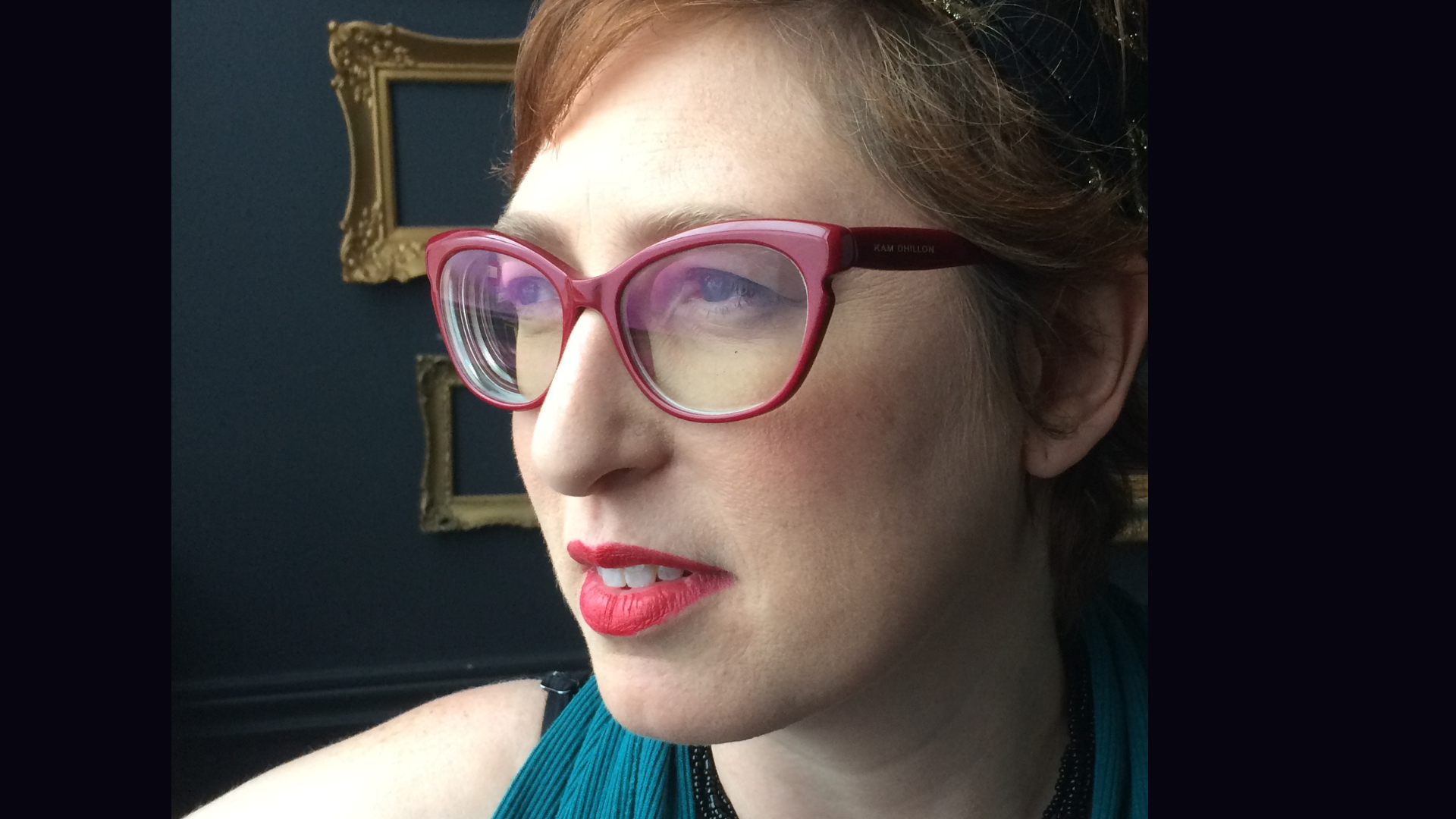 A photo of annie matan, a white woman in mauve glasses, wearing red lipstick and wearing short reddish hair.