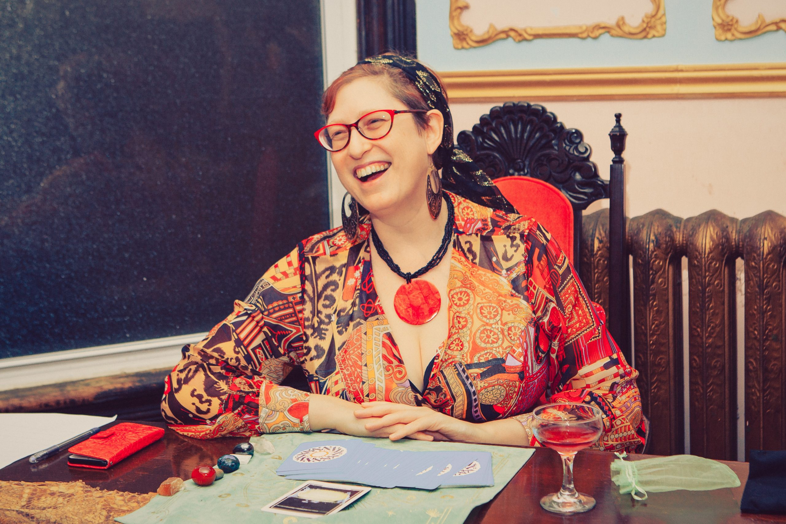 A white woman wearing mauve glasses and bright red dress with patter smiling with hands folded in front of her on the table