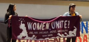 mage of two women holding a banner that reads WOMEN UNITE at IWD 2020 Rally