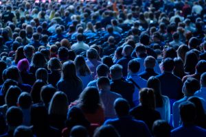 blue tinted image of a crowd of people at a business conference, facing towards the state