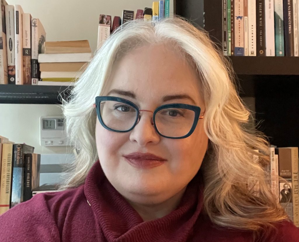 An image of a platinum blonde woman with blue glasses wearing a red turtle neck sweater. Book case behind her.