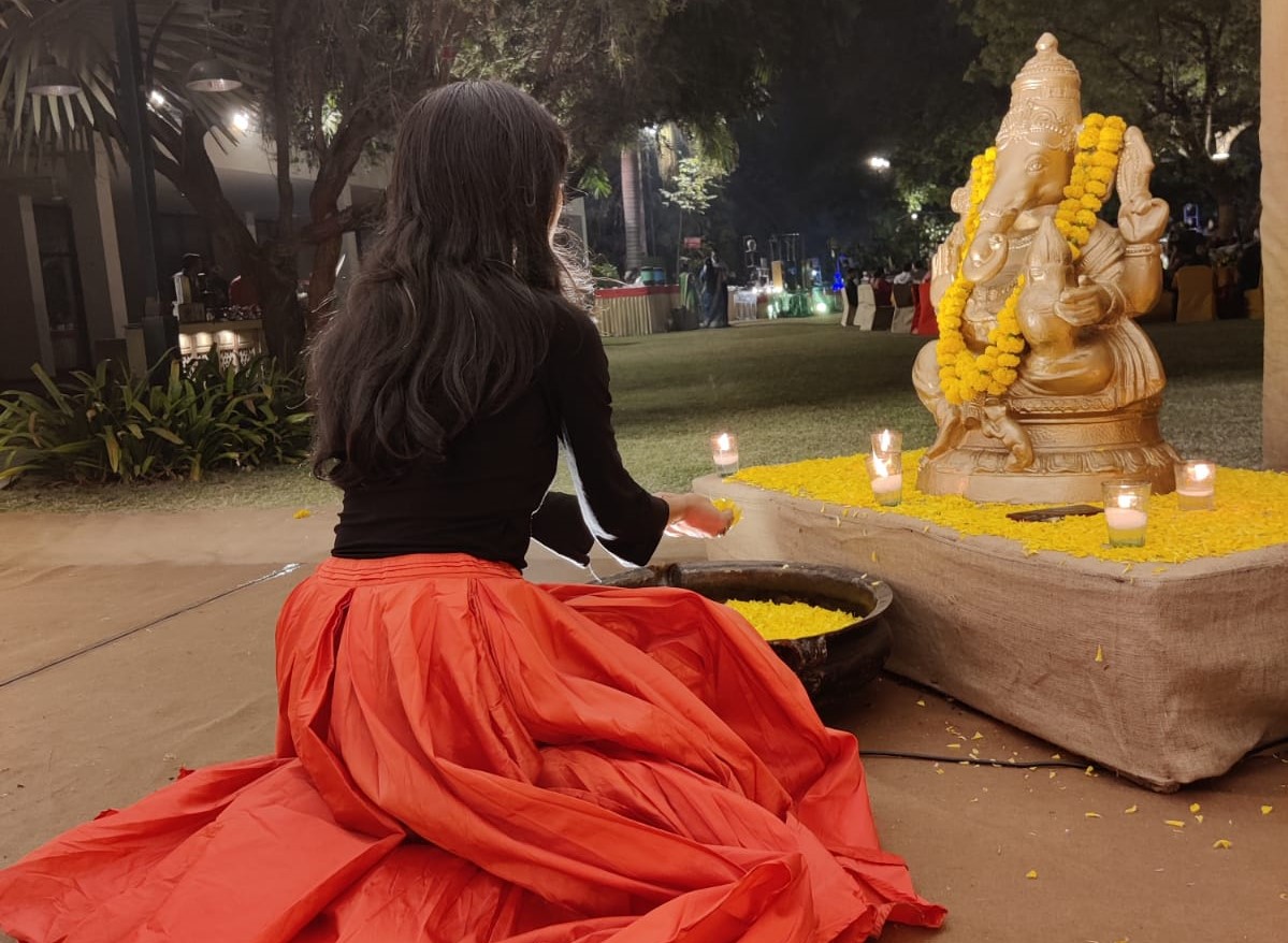 An picture of an indian woman kneeling before the God Ganesha. Whe is wearing a red skirt and black top.