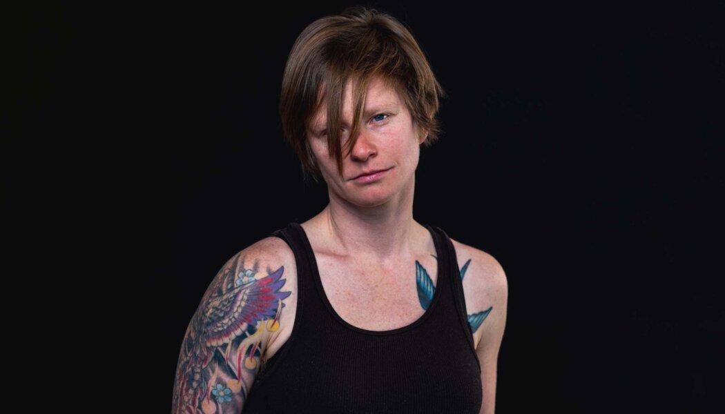 image of a white woman in a black tank top. She has tatoos on her left arm and shoulder. Short hair.