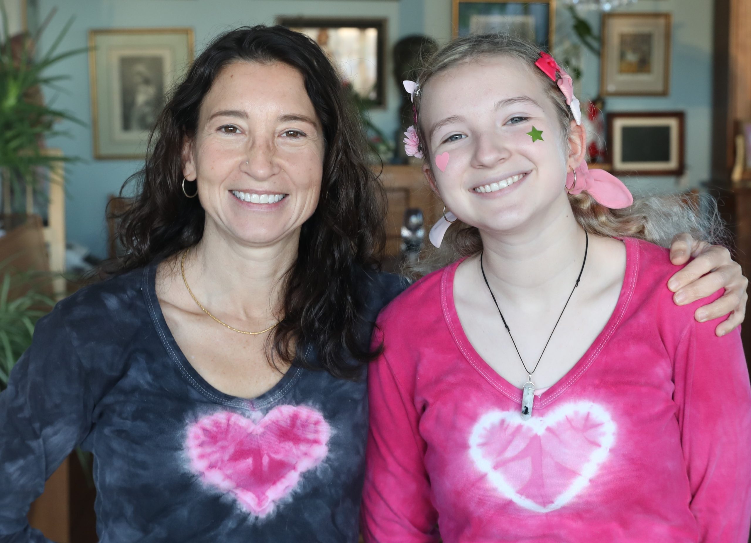 Image of mother and daughter. Mother wearing grey tshirt with a pink heart on the front. The daughter wearing pink tshirt with white heart. Mother has arm around the daughter.