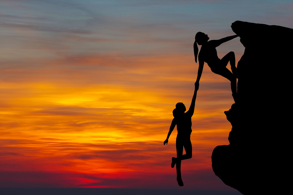 Photo of woman helping another women climb a cliff. Background is a sunset.