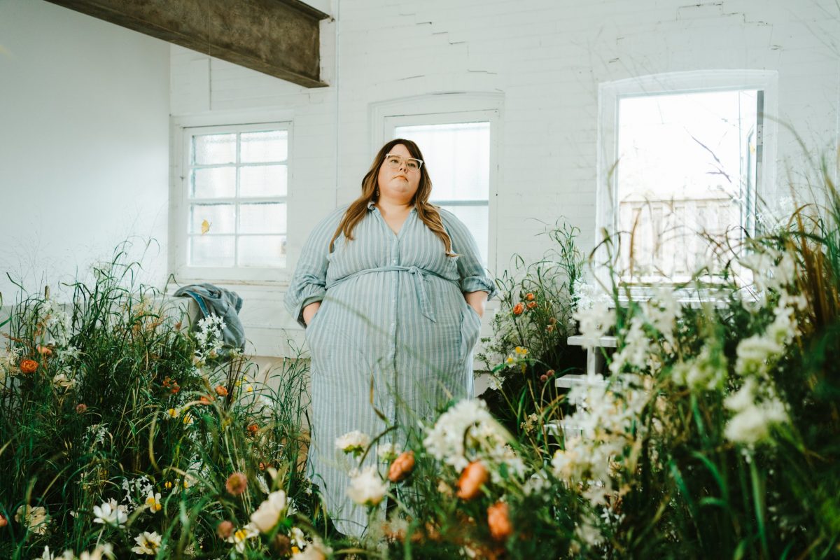 A woman, Cass Rudolph, with long brown hair wearing a blue stripped dress, standing in an indoor garden. White brick walls behind her.